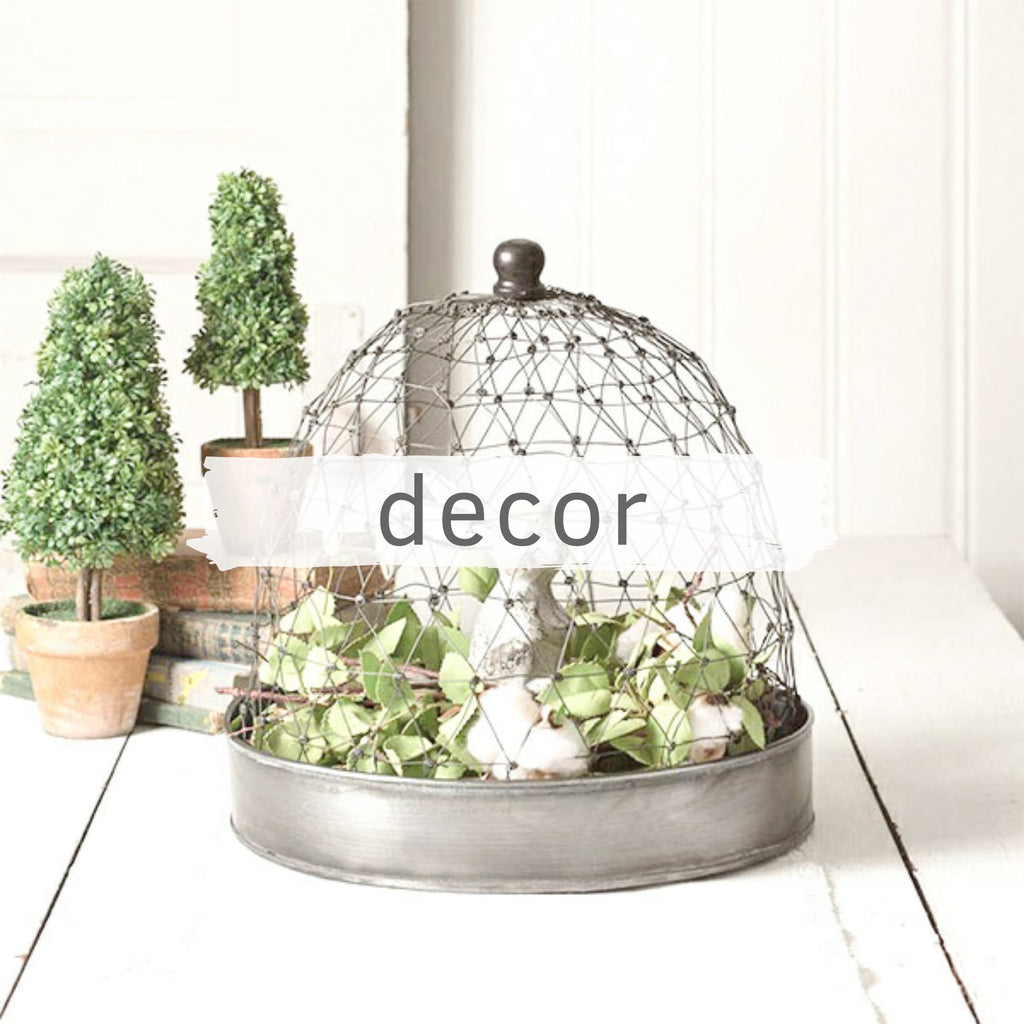 Galvanized, round tray with bunny figure and greenery and a wire cloche on top sitting next to two boxwood topiaries on a table. 
