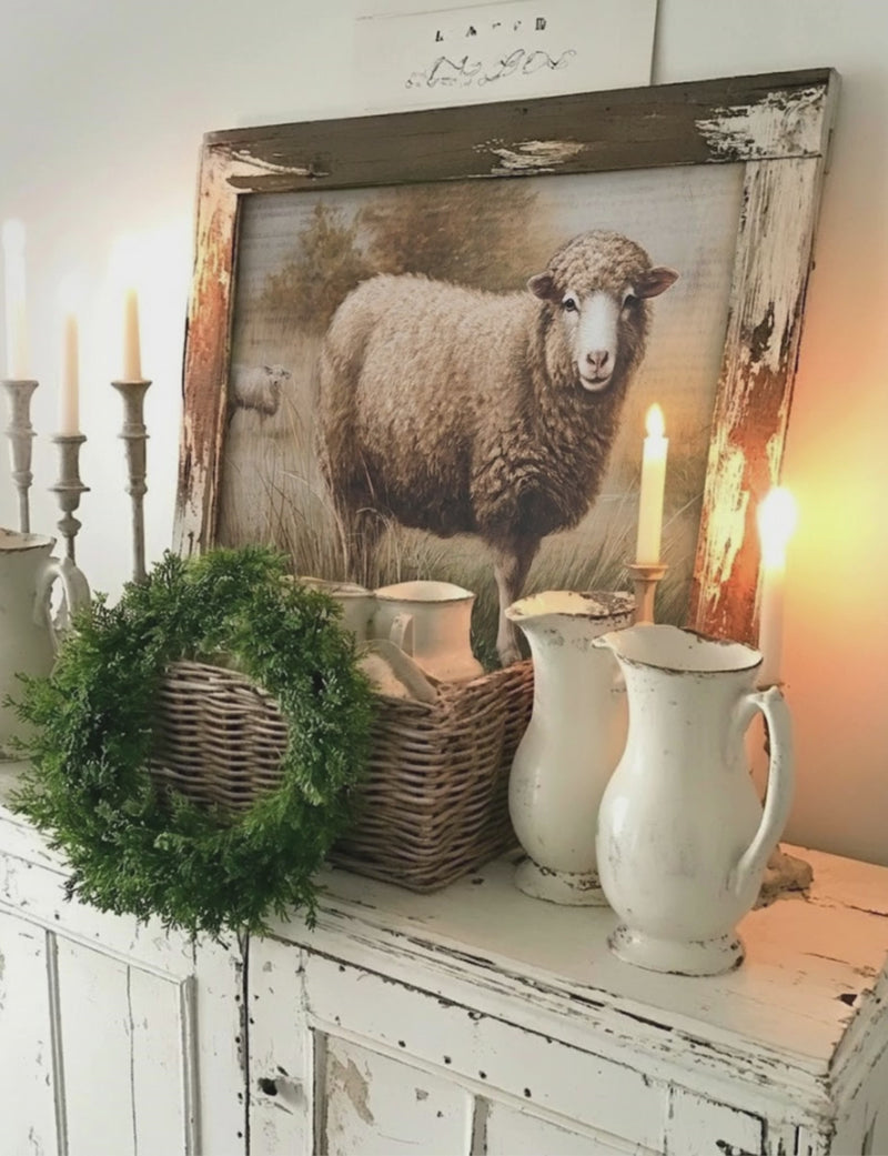 vignette farmhouse style with rustic framed sheep print, chippy pitchers, stacked dishes in basket and wreath